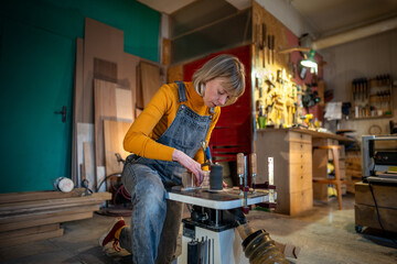 Focused woman joiner works in joinery workshop. Pedant female sanding, turning wooden knife blanks on a lathe. Small family business, furniture workshop, production of customised wooden tableware. - obrazy, fototapety, plakaty