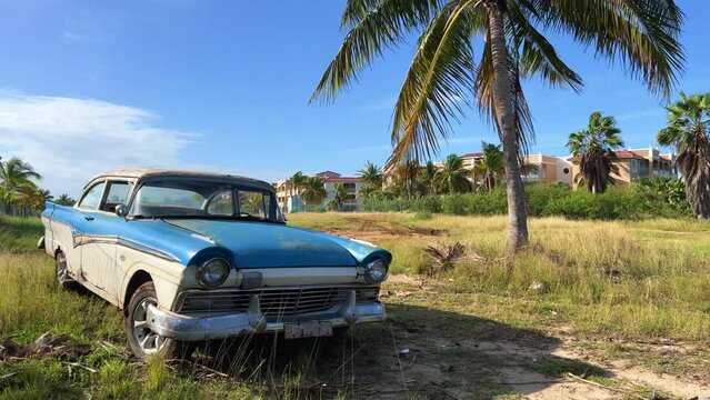VARADERO, CUBA - DECEMBER 2, 2024: An old American car standing under a palm tree, illuminated by the sun. A retro car. Blue classic American car on the beach. 4К