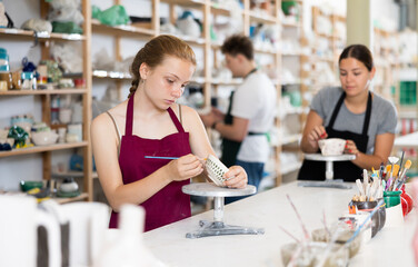 Girl teenager student in apron trained to paint ceramic cup in workshop