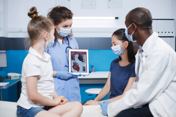 Fototapeta premium Black doctor shows cardiology image on digital tablet, pediatrician explains cardiovascular diagnosis to child and parent. Heart anatomy picture and heart disease problems presented by pediatrician.