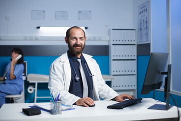 Middle aged doctor smiling at his desk in a medical office. Caucasian male physician appears to be...