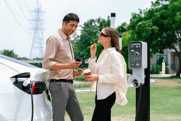 Young couple with coffee pay for electricity while recharge EV car battery at charging station...