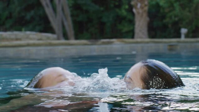 Two women playfully emerge from a swimming pool and push each other with a smile