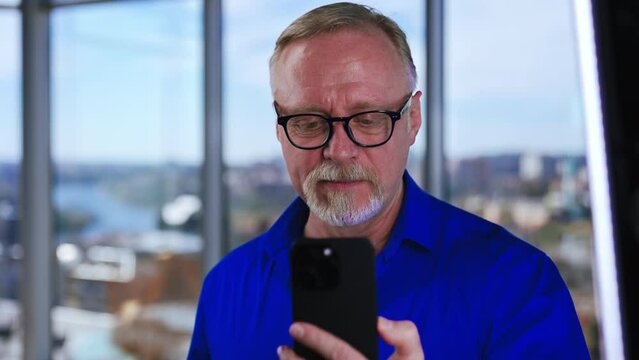 Cropped image of a mature man wearing blue shirt and glasses. Man looks at smartphone grimacing. Close up.
