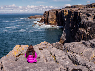 Teenager girl enjoys stunning view of mini cliffs in county Clare, Ireland. Warm sunny day. Travel and tourism. Irish landscape. Model with long hair, red jacket, slim body type. - obrazy, fototapety, plakaty