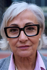 Vertical portrait of mature Caucasian business woman with glasses and gray hair in formal suit smiling looking to the camera outdoor. Female senior looks serious and confident