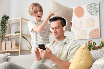 Young man in headphones using mobile phone and his naughty little son with pillow at home
