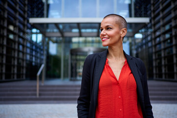 Modern stylish Caucasian adult business woman dressed in formal suit standing in work place outdoor. Strong and empowered female happy with shaved head looking smiling to the side. Copy space