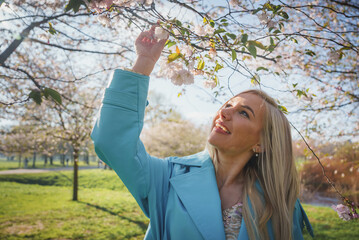 Summer fashion portrait of pretty blonde woman posing on amazing blooming Sakura tree background. Happy travel woman smile near a sakura cherry blossoms tree