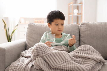 Ill little Asian boy with glass of water and pill sitting at home - obrazy, fototapety, plakaty