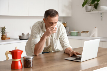 Mature man with laptop drinking hot coffee at table in kitchen