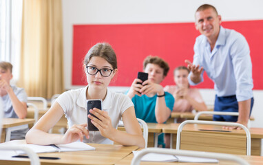 Concentrated teenage girl with group of fellow students sitting at lesson in classroom, using...