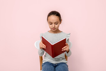 Little girl reading book while sitting on chair against pink background