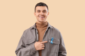 Young man pointing at blue ribbon on beige background. Prostate cancer awareness concept