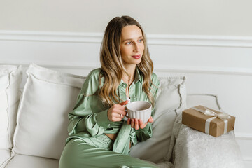 Young beautiful blonde woman with cup of coffee sitting home relaxing by the window in living room wearing pajamas. Lazy day off concept.