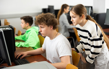Positive teen girl student talking to concentrated teenager studying in college library computer...