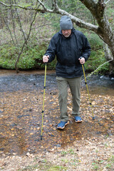 Senior hiker crossing creek in cold weather gear with hiking poles.