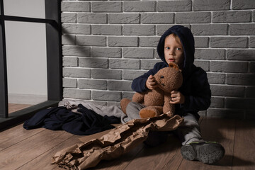 Homeless little boy with toy bear sitting near grey brick wall