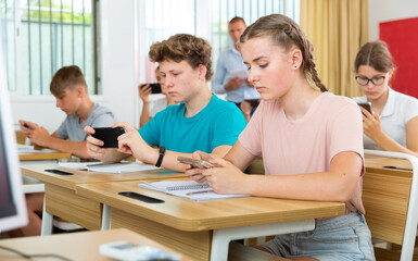 Group of modern teenagers sitting at lesson in classroom, using mobile phones..