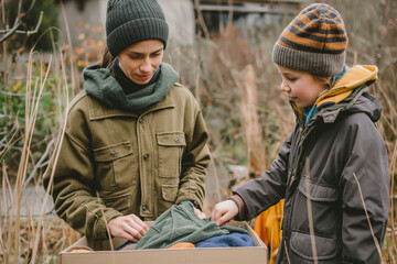 Fototapeta premium Woman and child sorting clothes and packing into cardboard box. Donations for charity, help low income families, declutter home, sell online, moving moving into new home, recycling, sustainable living