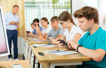 Fototapeta premium Pupils sitting in class room and watching in their smartphones.