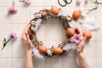 Woman making Easter wreath with eggs, feathers and flowers on white tile background