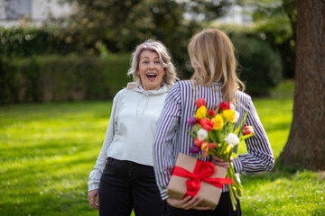 Daughter surprises mother with hidden gift and flowers behind back.