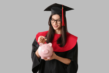 Beautiful young happy female graduate with piggy bank and coin on grey background. Tuition fees...