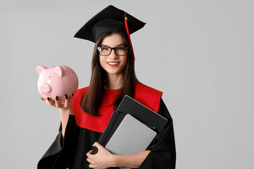 Beautiful young happy female graduate with piggy bank and notebooks on grey background. Tuition...