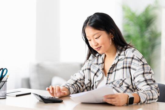 Young chinese woman preparing home budget using calculator sitting in the living room