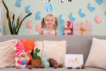 Cute little girl in bunny ears with chocolate Easter eggs and toys on sofa at home