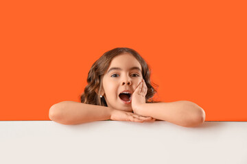 Shocked little girl with blank poster on orange background