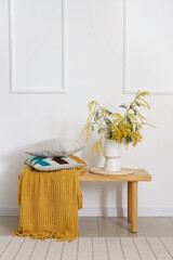 Wooden table with soft cushions, yellow blanket and mimosa flowers near white wall