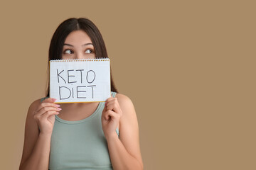 Beautiful young thoughtful Asian woman holding notebook with text KETO DIET on brown background