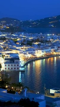View of Mykonos town with famous windmills, and port with boats and cruise ship illuminated in the evening blue hour. Mykonos, Cyclades islands, Greece. With camera panning