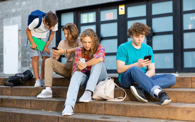 Teenager girl sitting on stairs beside school building and using smartphone. Her classmate sitting...