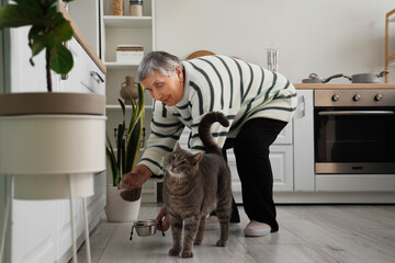 Senior woman feeding cute cat in kitchen