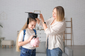Mother putting mortar board on her daughter with piggy bank at home. Education savings concept