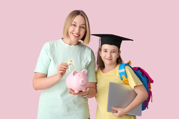 Mother and her daughter in mortar board with laptop putting money into piggy bank on pink...