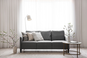 Grey sofa and vase with blooming branches on coffee table in interior of light living room