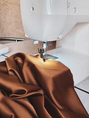 The image shows a close-up of a sewing machine stitching brown silk fabric. The image is well-lit,...