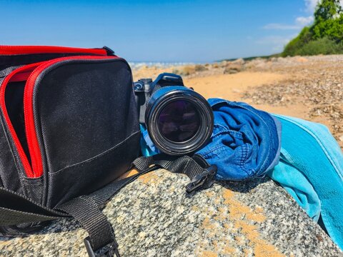 Camera and beach essentials on a rock by the sea with blue towel and jeans. Summer travel photography gear in nature for relaxation. Tranquil getaway with blue sky and peaceful sea view.