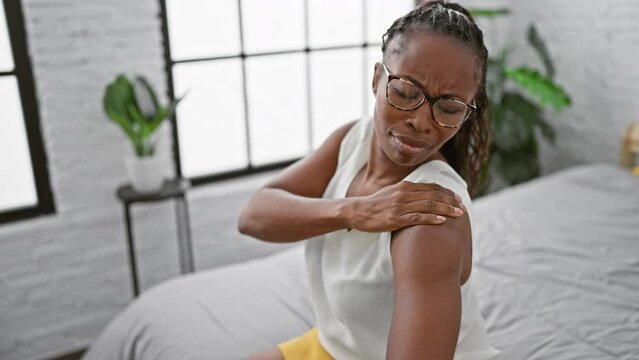 A woman experiences shoulder pain in a bright bedroom setting, expressing discomfort.