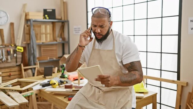 A focused man with a beard in a workshop, talking on the phone and holding a notebook.