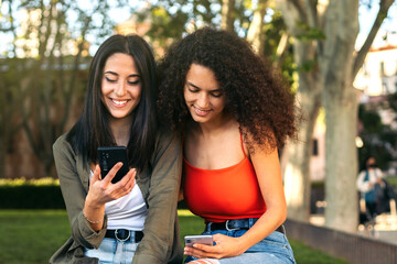 two young women sitting using her mobiles in a park