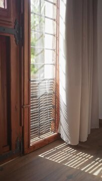 Sunlight filters through a wooden window and blinds, casting shadows on a curtain and hardwood floor in a cozy, serene room.