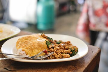 A plate of pad kra pao (holy basil pork mince stir-fry) with omelette and mixed grain rice at a street food stall near the Grand Palace in Phra Nakhon, Bangkok, Thailand - obrazy, fototapety, plakaty