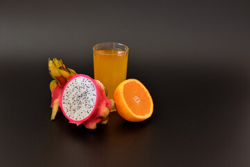 A tall glass of a mixture of tropical fruit juices on a black background, next to the halves of a ripe orange and pitaya.