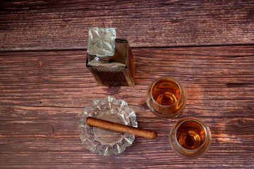 A crystal decanter, two glasses of cognac and a crystal ashtray with a Cuban cigar on a dark wooden table.