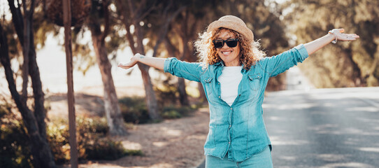 Portrait of wonderful white female model with curly blonde hair expressing energy on a good day in Europe. Beautiful curly woman smiling and walking with trees behind her. - obrazy, fototapety, plakaty